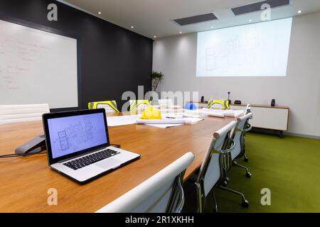 Laptop with blueprint, hardhat on conference table surrounded with empty chairs in meeting room Stock Photo