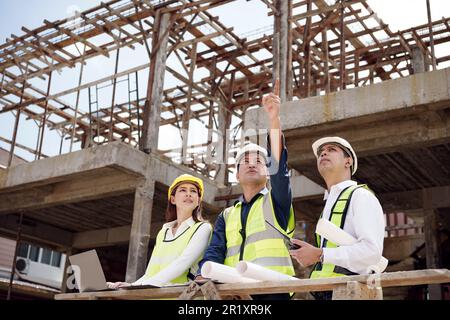 Asian Architector and engineers team with draft plan of building and tablet talking constructing site. Construction manager and engineer working on bu Stock Photo