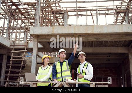 Asian Architector and engineers team with draft plan of building and tablet talking constructing site. Construction manager and engineer working on bu Stock Photo