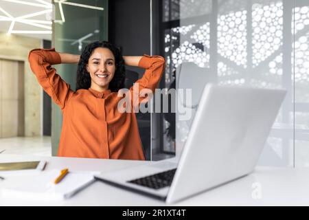Portrait of successful Hispanic businesswoman, female worker at workplace resting after successful completion of work, woman smiling and looking at camera, satisfied with herself, hands behind head. Stock Photo