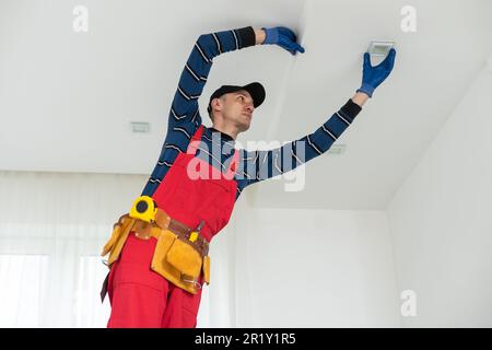 man plasterer construction worker at work, takes plaster from bucket and puts it on trowel to plastering the wall, wears helmet inside the building Stock Photo