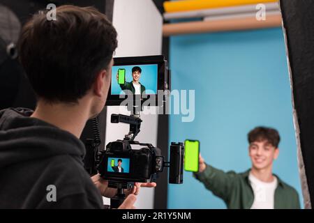 View of a professional photographer shooting a young man showing smartphone with green screen, blue background Stock Photo