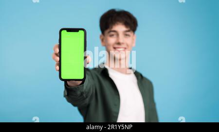 Young man smiling and showing his phone with green screen into the camera on a blue background Stock Photo
