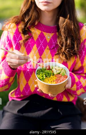Close view of a woman eating a poke bowl with vegetables, fruits and fish using chopsticks Stock Photo