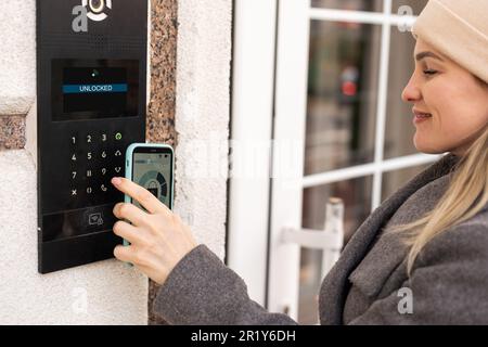 Young stylish woman getting access to the building by attaching smartphone to intercom. Concept of modern security technologies for access and smart Stock Photo