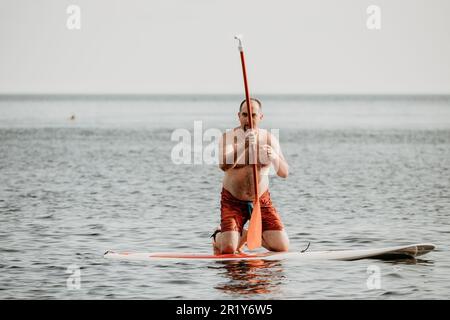 Active mature male paddler with his paddleboard and paddle on a sea at summer. Happy senior man stands with a SUP board. Stand up paddle boarding - Stock Photo