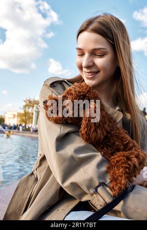 A young smiling blonde girl in a stylish beige raincoat holds and hugs her pet toy poodle in her arms. The girl and the dog are resting in the park. S Stock Photo