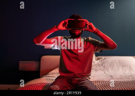 Black man using virtual reality headset at home with neon light, sit on bed at night. VR addiction. Stock Photo
