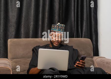 Good looking businessman in Africa attire seated working on his laptop and using his mobile phone to perform business transactions Stock Photo
