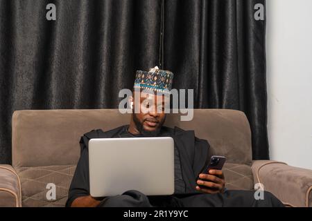 Good looking businessman in Africa attire seated working on his laptop and using his mobile phone to perform business transactions Stock Photo