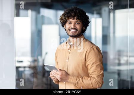 Portrait of young programmer with tablet computer inside office, hispanic man with curly hair smiling and looking at camera, man at workplace testing online applications standing near window Stock Photo