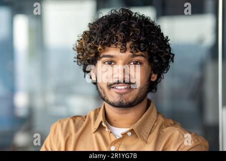 Young smiling indian programmer close up smiling and looking at camera, portrait of man with curly hair and shirt inside office at work, businessman entrepreneur with beard working on project Stock Photo