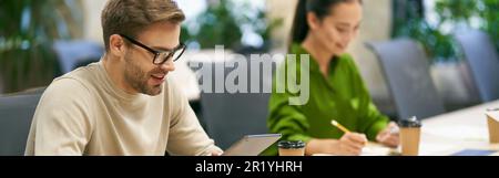 Working together. Young smiling caucasian man wearing eyeglasses using digital tablet while sitting at desk with his female colleague in the modern Stock Photo