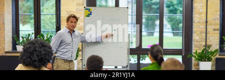 Male business coach or speaker pointing at flip chart, giving presentation to audience, standing against multiracial group of people sitting at desks Stock Photo