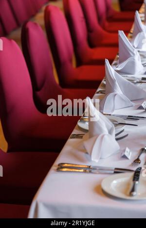 A long dining table set out for a large party with red chairs and white table place settings. Stock Photo