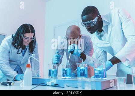A diverse team of scientists is working in a chemistry lab. One of them is using a dropper to add liquids to vials in a magnetic stirrer, creating bub Stock Photo