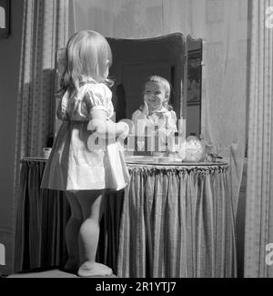 In the 1950s. A little girl is standing in front of her mothers makeup table and mirror and puts on some kind of beauty cream on her face, looking happy. Sweden 1951 Conard ref 1708 Stock Photo