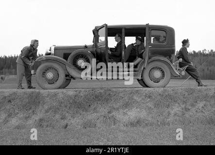 The old car. A family out driving with their vintage car looks as if it's stopped and they are trying to start it again. The early cars could also be started with a crank, if the battery had ran out of power. A woman is seen using it in front of the car while the man is behind the wheel. By the look of the passangers faces and body language this may have been a long ongoing attempt.   Sweden 1952. Kristoffersson ref 12K-24 Stock Photo