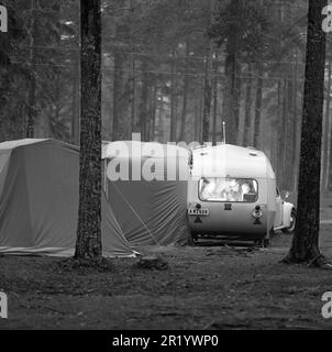 1960s camping. A family is enjoying their holiday and the practial camping life in their caravan. It's evening and they are having dinner inside the caravan. The special tent used in combination with the caravan trailer is part of the camping gear. Sweden 1962 Ref BV60 Stock Photo
