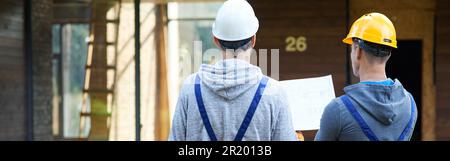 Plan it. Rear view of two builders standing outdoors with an open blueprint discussing the interior of a half completed cottage house Stock Photo