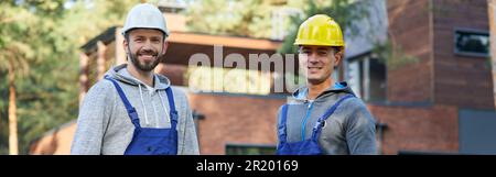 Rely on us. Two positive handsome young male engineers in hard hats smiling at camera, posing outdoors with toolbox and suitcase with tools while Stock Photo