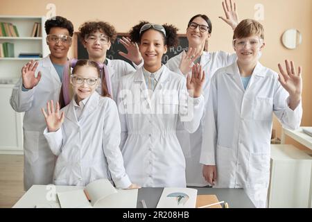 Diverse group of happy teenagers wearing lab coats in school and waving at camera standing together Stock Photo