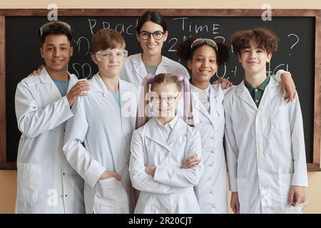 Diverse group of children wearing lab coats posing with teacher in science class at school and smiling at camera Stock Photo