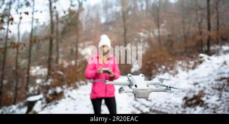 Woman with a controller in her hands navigating a drone floating in the air in front of her. Outside in winter forest. Focus on the drone. Stock Photo