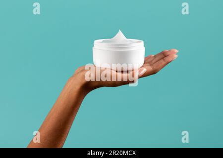 Unrecognizable African American female showing white plastic jar with body cream against turquoise background Stock Photo