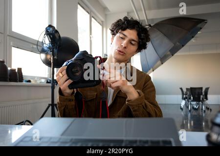 European male photographer using modern photo camera while working on laptop at workplace in studio Stock Photo