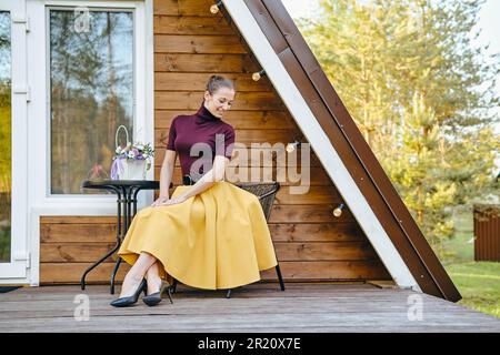 Cute shy woman in long skirt and blouse sits on terrace of wooden cabin Stock Photo