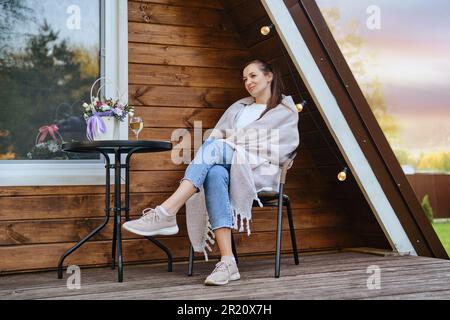 On warm evening, woman wraps herself in cozy blanket and rests on veranda of tiny house. Concept of small modern cabins for rest and escape to nature Stock Photo