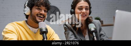 excited and curly indian man in yellow jumper with charming brunette woman in grey blazer talking in professional microphones while working and record Stock Photo