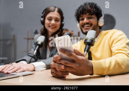 cheerful indian radio host in headphones and yellow blazer holding smartphone near brunette colleague using laptop close to professional microphones i Stock Photo