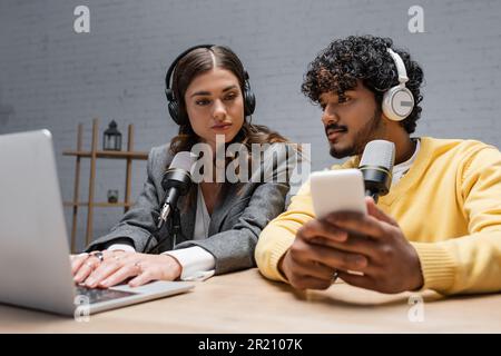 charming brunette podcaster in headphones and grey blazer using laptop near young indian radio host holding blurred mobile phone while working in radi Stock Photo