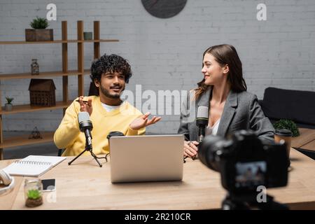 positive indian man in yellow jumper gesturing near microphones, laptop, notebook and smartphone while talking to smiling colleague sitting in blazer Stock Photo
