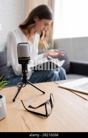 selective focus of table with laptop and eyeglasses and professional microphone near podcaster sitting on couch and working with notebook on blurred b Stock Photo