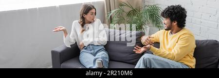 emotional brunette woman in white blouse gesturing while talking to young and curly indian man in yellow jumper sitting on sofa near green plant again Stock Photo