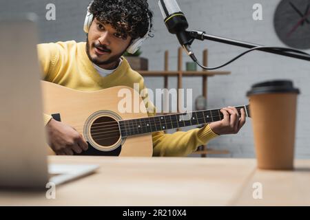 cheerful indian podcaster in headphones and yellow jumper playing acoustic guitar near microphone and blurred laptop with coffee to go in paper cup on Stock Photo