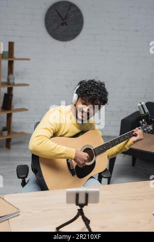 joyful indian musician in headphones and yellow jumper playing acoustic guitar near blurred mobile phone on tripod and professional microphone while r Stock Photo