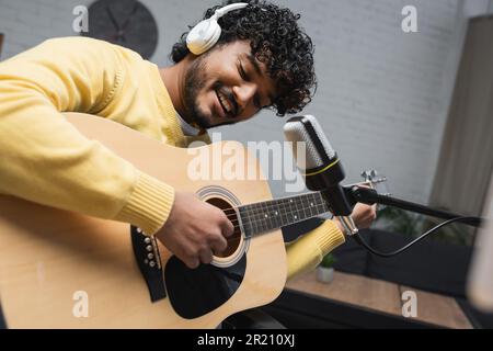 Positive and young indian podcaster in headphones playing acoustic guitar near microphone on stand during stream in podcast studio,stock image Stock Photo