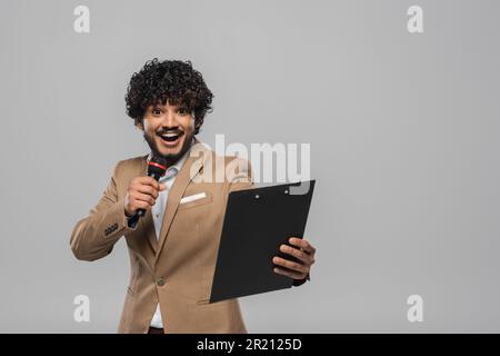 Cheerful and young indian host of event with curly hair standing in formal wear talking at microphone and holding clipboard during party isolated on g Stock Photo