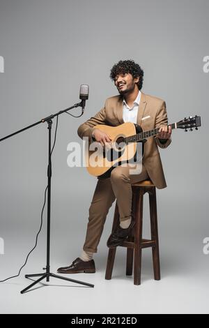 Smiling indian event host in formal wear playing acoustic guitar and singing in microphone in stand while sitting on chair on grey background,stock im Stock Photo