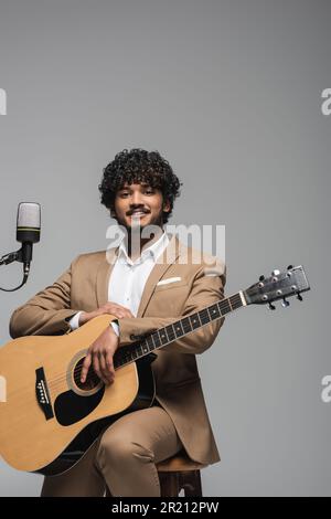 Young smiling indian man in formal wear holding acoustic guitar and looking at camera while sitting on chair near microphone isolated on grey,stock im Stock Photo