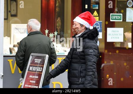 A shopper wearing a mask and a Santa hat walks along Hornchurch high street in the London Borough of Havering amid the COVID-19 coronavirus pandemic as a new COVID-19 variant known as B.1.1.529 or Omicron continues to spread 2021. Stock Photo