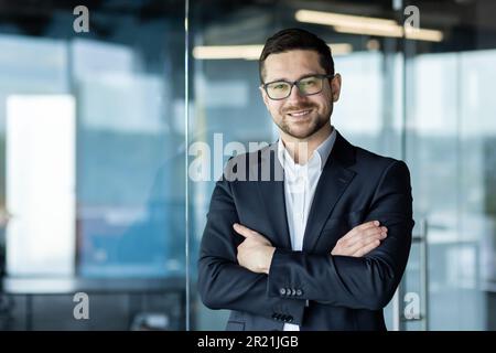 Portrait of a young self-confident male teacher, tutor, mentor standing in an office, campus in a suit and glasses, crossing his arms and looking at t Stock Photo