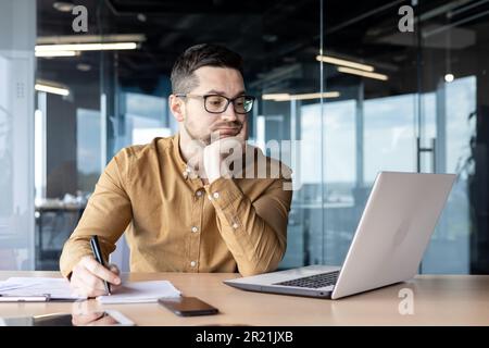 Tired and bored young man businessman, accountant, manager sitting at a table and working on a laptop and with documents. Propping his head with his h Stock Photo