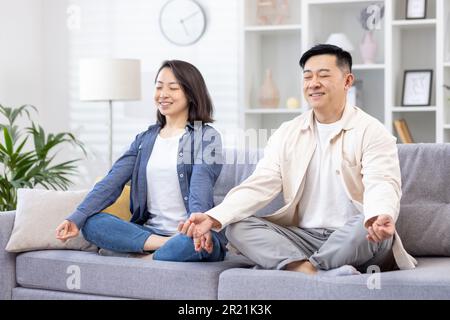 Young Asian young family, couple, man and woman spend time together, rest, do yoga. They are sitting on the sofa in the lotus position with their eyes closed, smiling, relaxing. Stock Photo