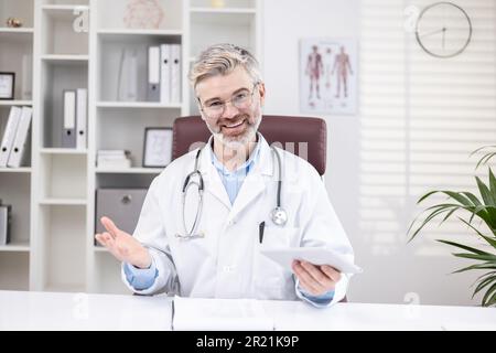 Portrait of a smiling senior male doctor who remotely consults patients online, sits in the office at the table and talks to the camera on a video call Stock Photo