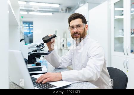 Portrait of a young male student, an intern sitting at a laboratory table, studying at a laptop and doing research through a microscope. Smiling looking at the camera. Stock Photo
